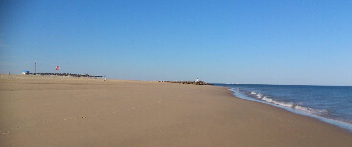 A wide sandy beach with gentle waves, a clear blue sky, and a lighthouse in the distance.