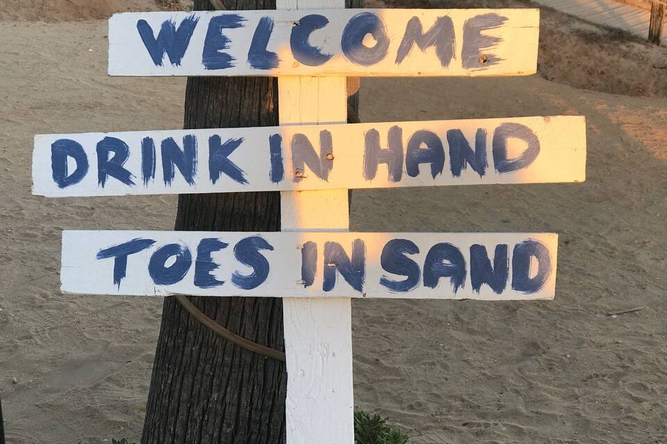 A wooden sign with 'WELCOME', 'DRINK IN HAND', and 'TOES INSAN' painted on it.