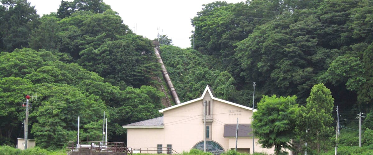 A small building with a sloped roof, surrounded by lush green trees and a stone embankment.