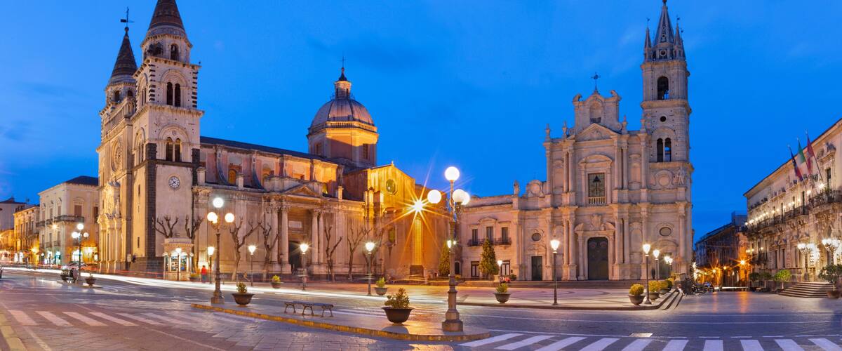 A historic square with ornate buildings, including a church with a dome and twin spires, under twilight sky.