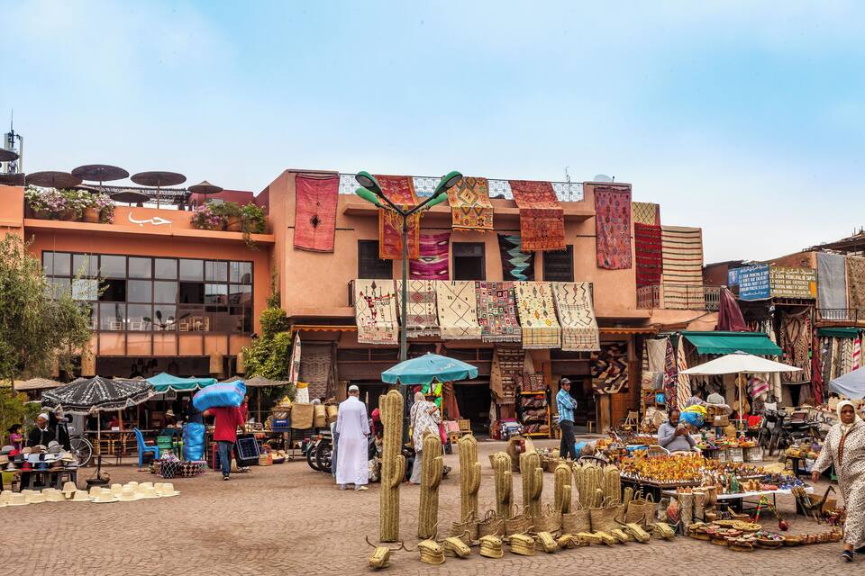 Un mercat animat amb tèxils de colors penjats als edificis, diferents parades que venen productes i persones passejant.