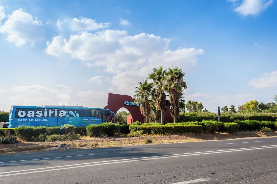 A blue bus with 'oasiria' and 'PARC AQUATIQUE' on it, a red building with 'Qasiria' on it, palm trees, and a road.