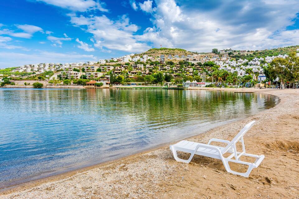 A beach with a white lounge chair, calm water, and a hillside town in the background.