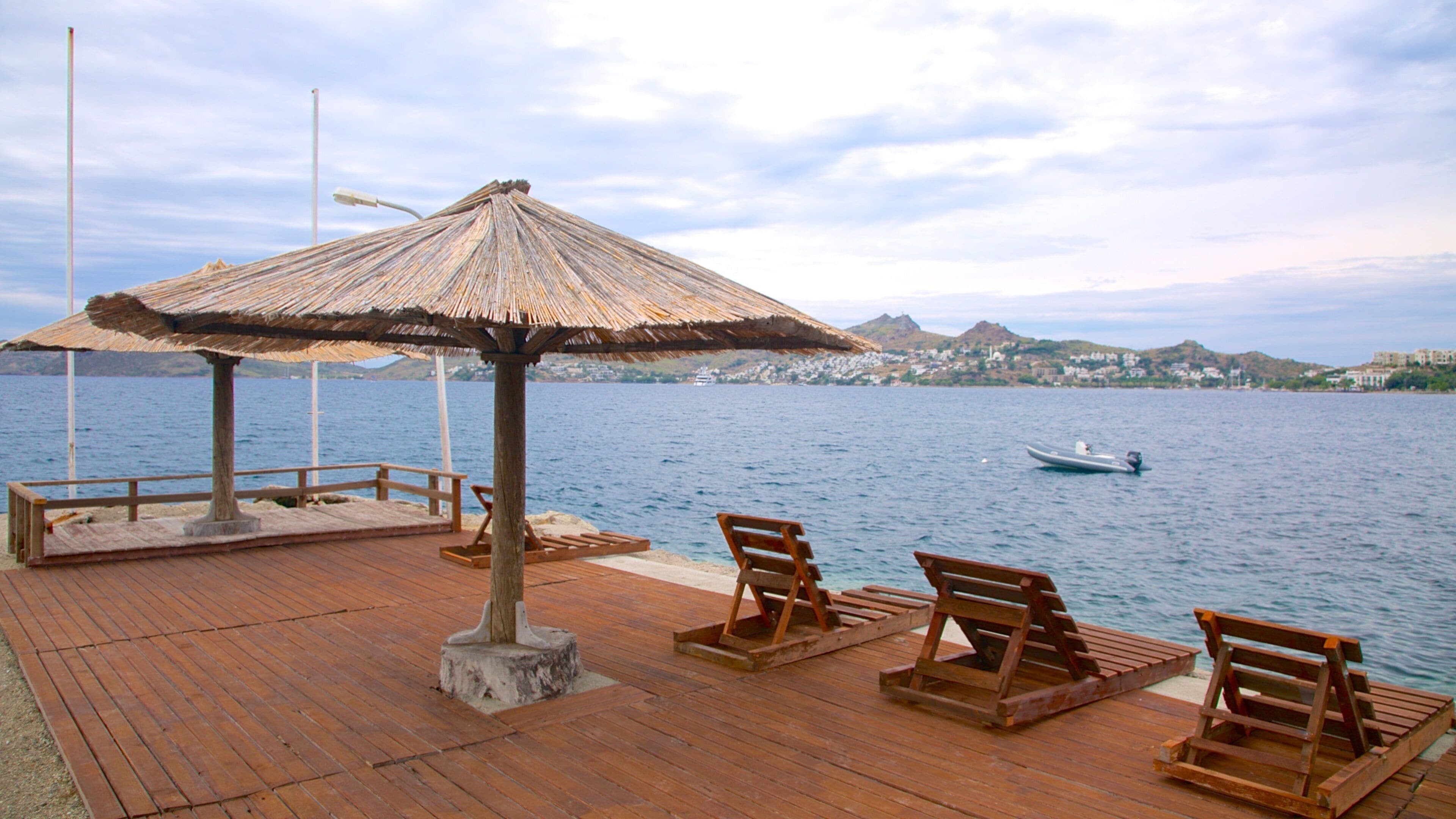 A wooden deck with a thatched umbrella and wooden chairs by a calm sea.