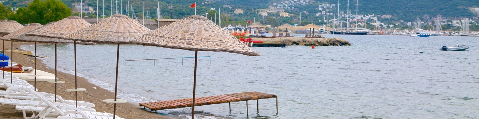 Una playa con sillas reclinables blancas y sombrillas de paja, un rompeolas rocoso y barcos al fondo.