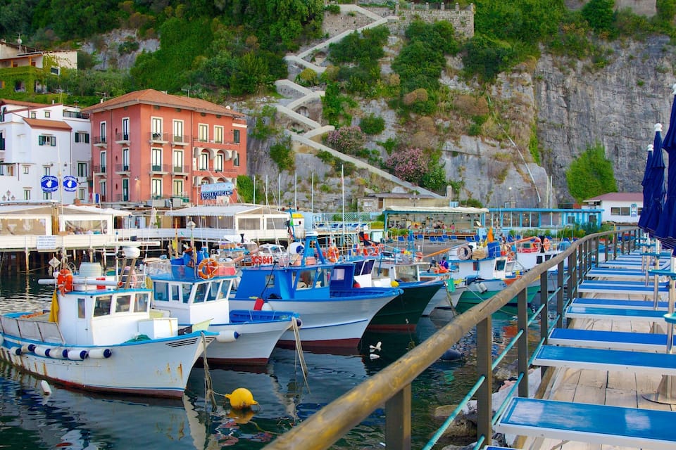 A harbor with boats docked, a pier with blue and white seating, and buildings with balconies overlooking the water.