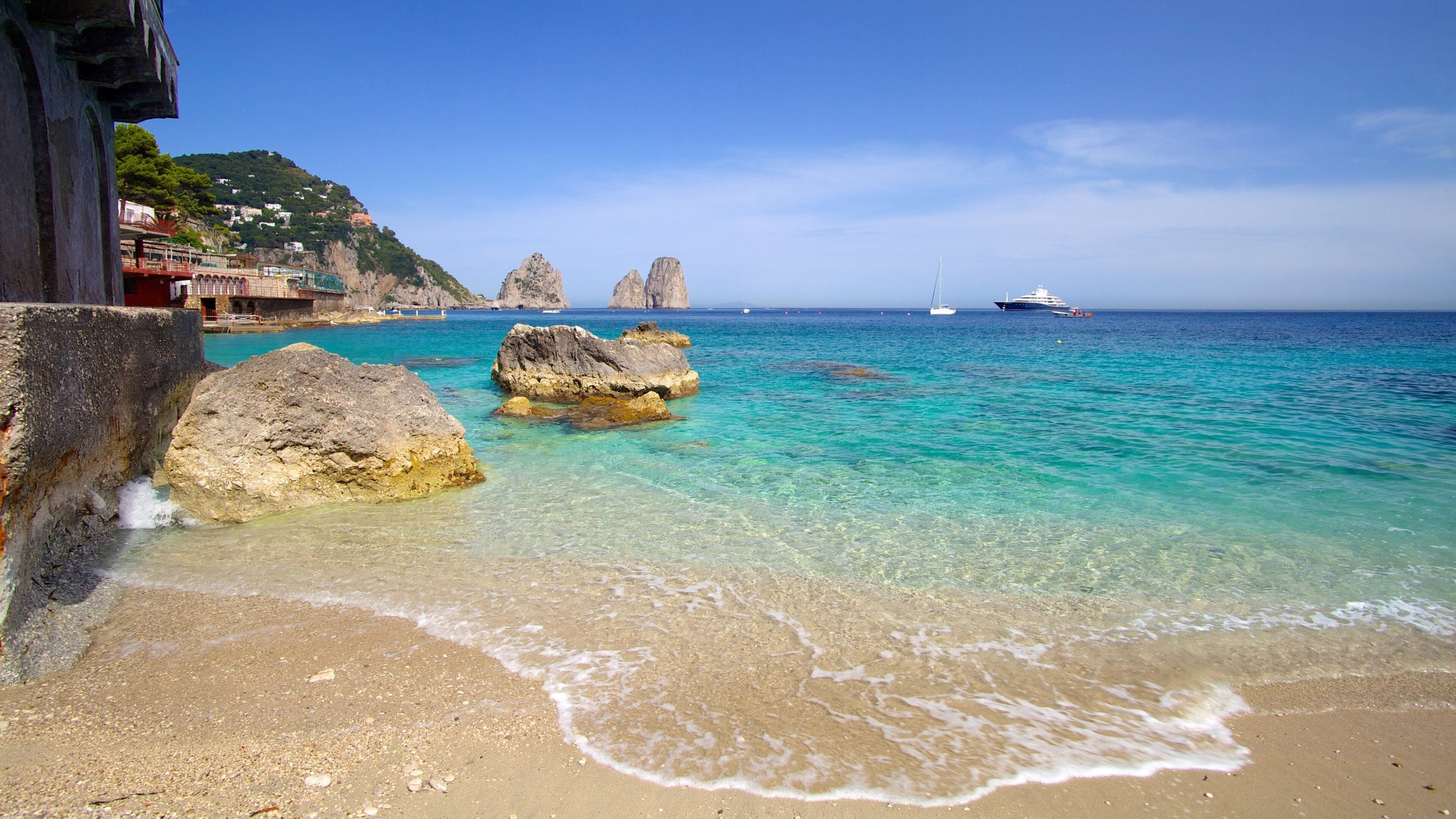 A beach with clear turquoise water, large rocks, and a distant sailboat.