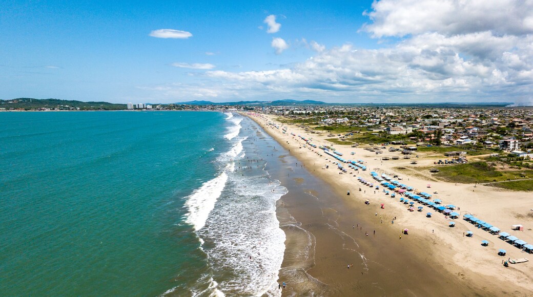 Aerial drone view of the beach, tourists, sea and waves splashing in the coast of the town Playas General Villamil, Ecuador. Sunny day. Beach houses and the horizon with some clouds in background.