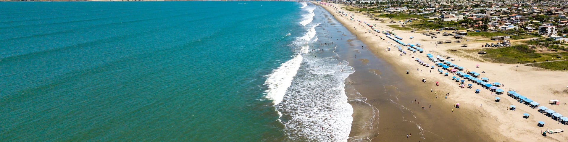 Aerial drone view of the beach, tourists, sea and waves splashing in the coast of the town Playas General Villamil, Ecuador. Sunny day. Beach houses and the horizon with some clouds in background.