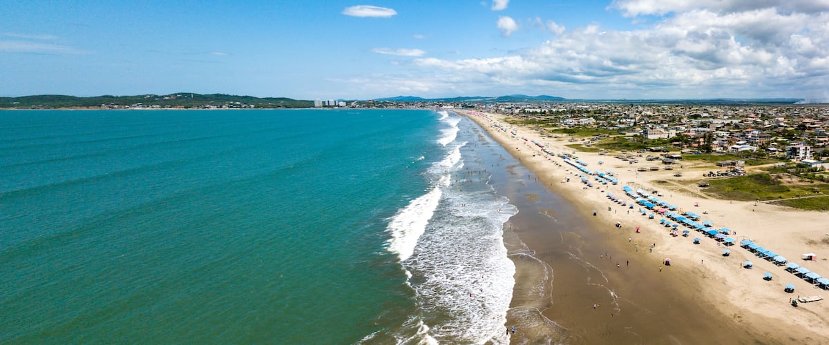 A coastal city with a long sandy beach, a clear blue sea, and distant hills.
