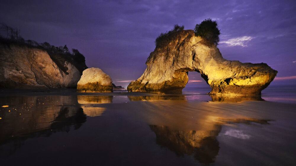 A rock arch formation at twilight with reflections in calm waters.