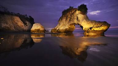 Formación rocosa en forma de arco al atardecer, con reflejos en aguas tranquilas.