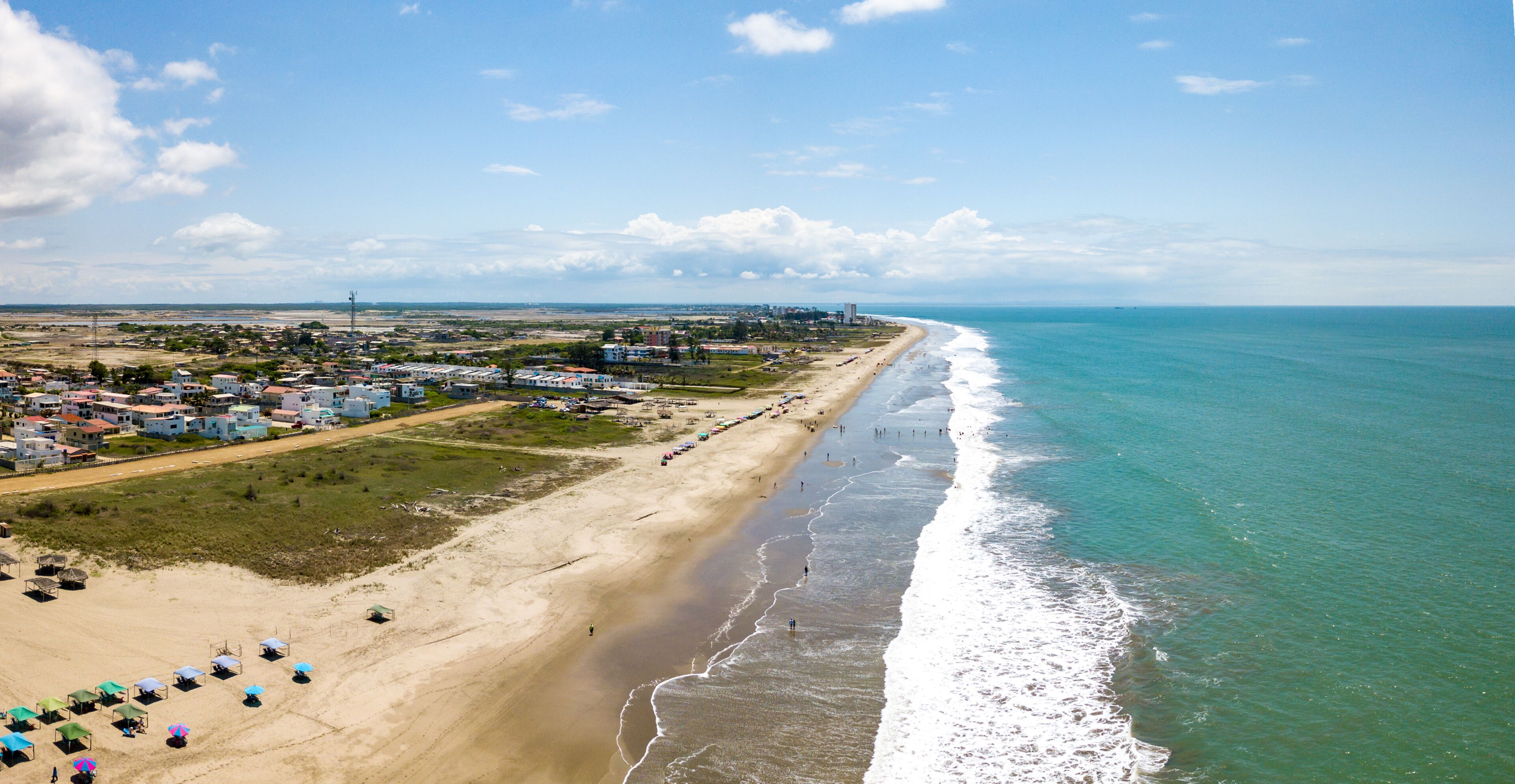 Aerial drone view of the beach, tourists, sea and waves splashing in the coast of the town Playas General Villamil, Ecuador. Sunny day. Beach houses and the horizon with some clouds in background.