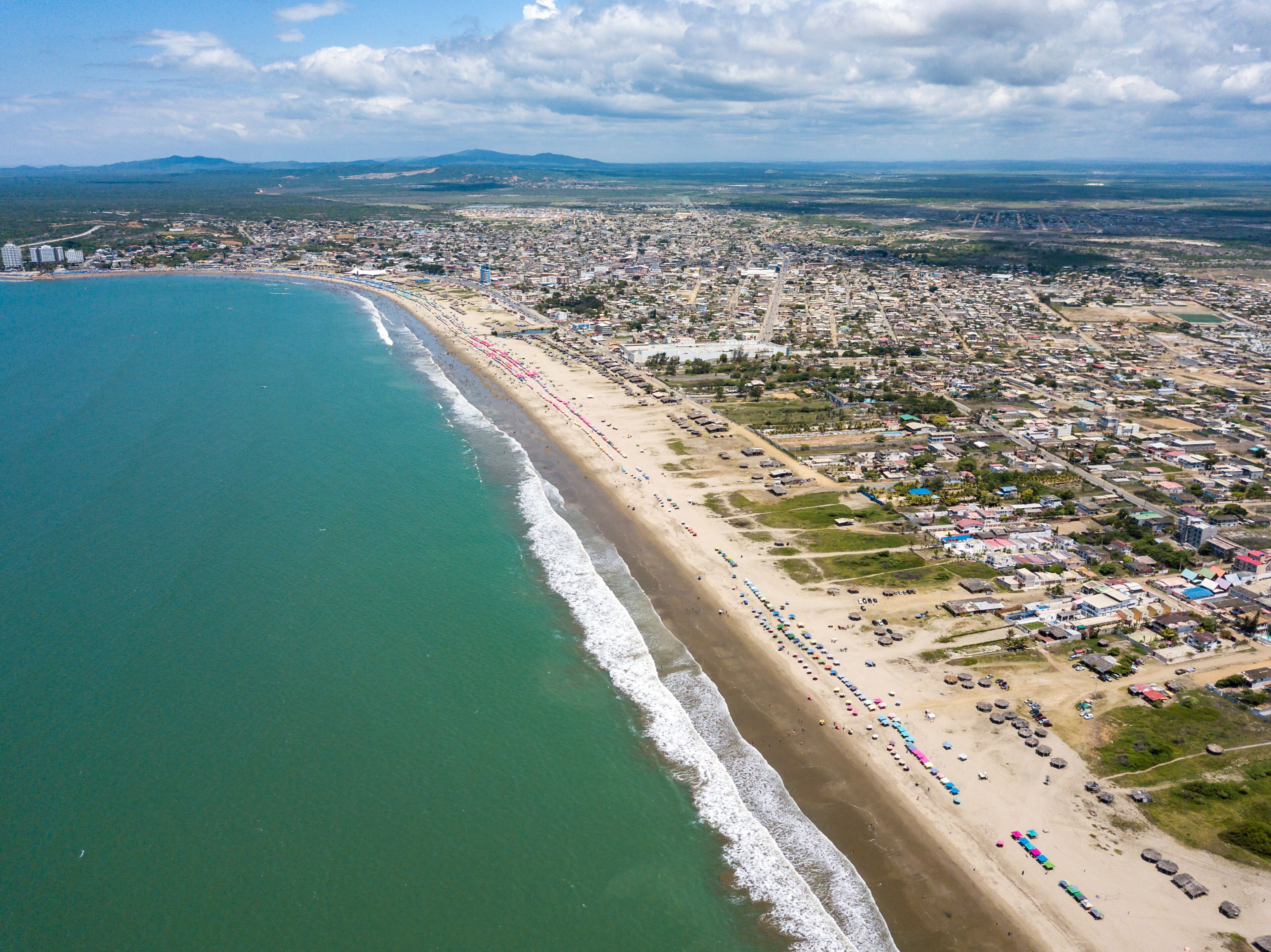 Aerial drone view of the beach, tourists, sea and waves splashing in the coast of the town Playas General Villamil, Ecuador. Sunny day. Beach houses and the horizon with some clouds in background.