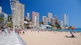 A beach with people sunbathing and walking, surrounded by tall buildings and palm trees.