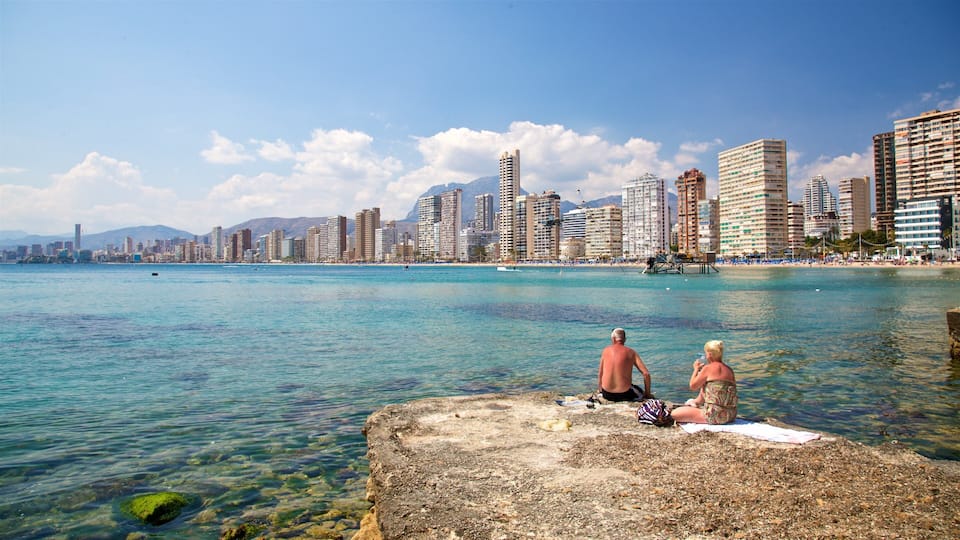 A coastal cityscape with clear blue waters, a rocky shoreline, and high-rise buildings in the background.