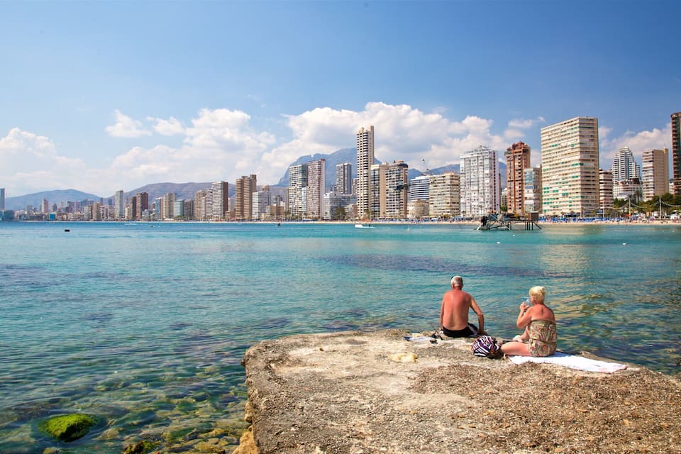 Paisagem costeira com águas azul-claras, um litoral rochoso e edifícios altos ao fundo.