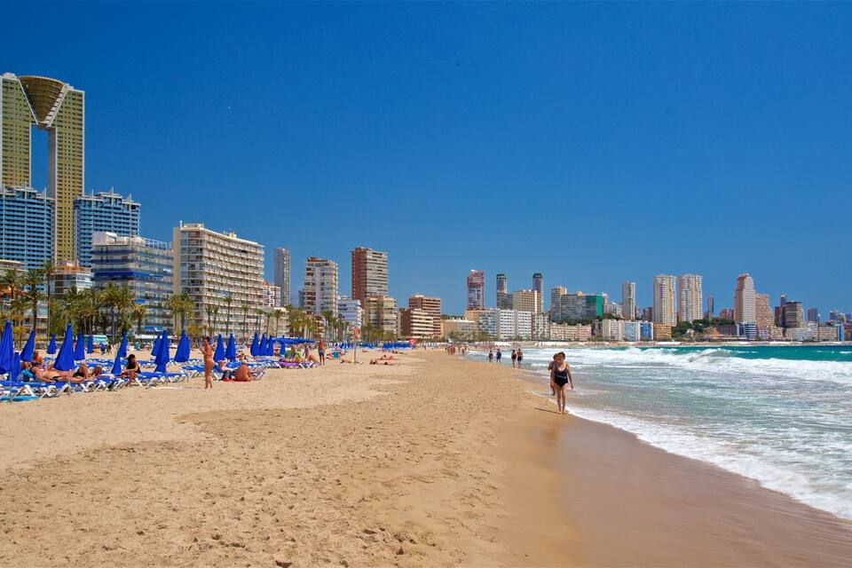 A beach with blue umbrellas and people sunbathing, a city skyline in the background, and waves crashing onto the shore.
