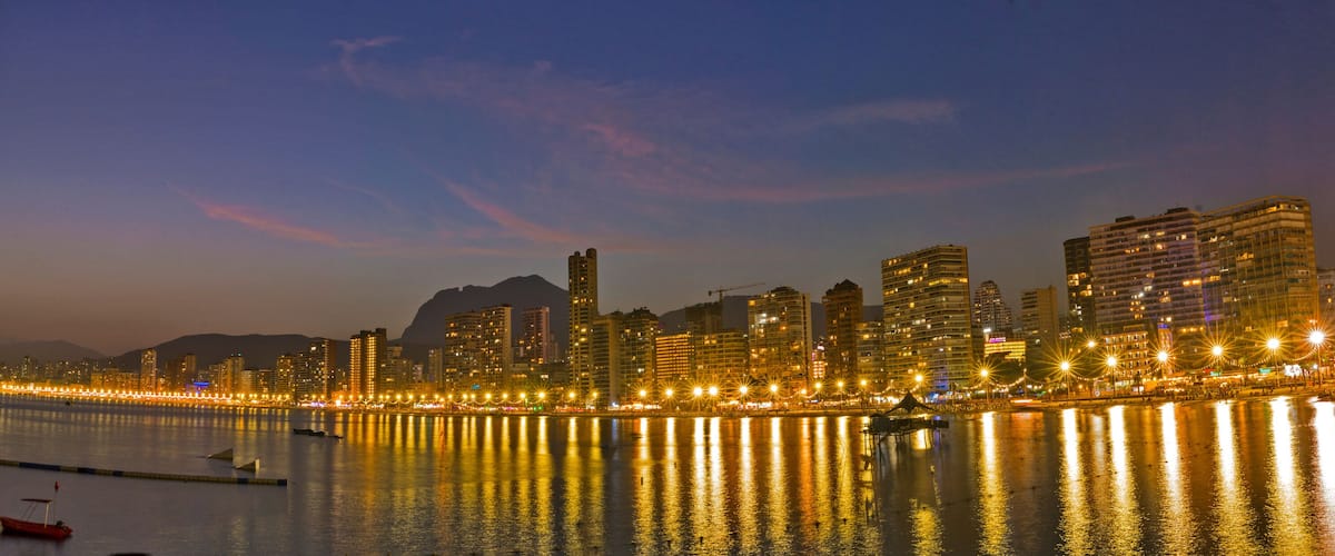 A city skyline at dusk with illuminated buildings reflecting on the water.