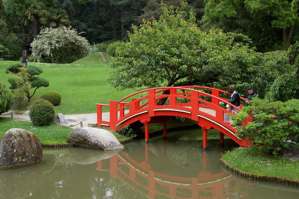 A red bridge over a pond in a lush garden with trees and rocks.