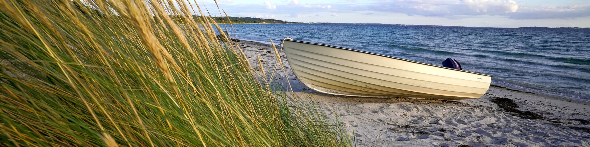 A boat on a sandy beach with tall grass in the foreground and a calm sea in the background.