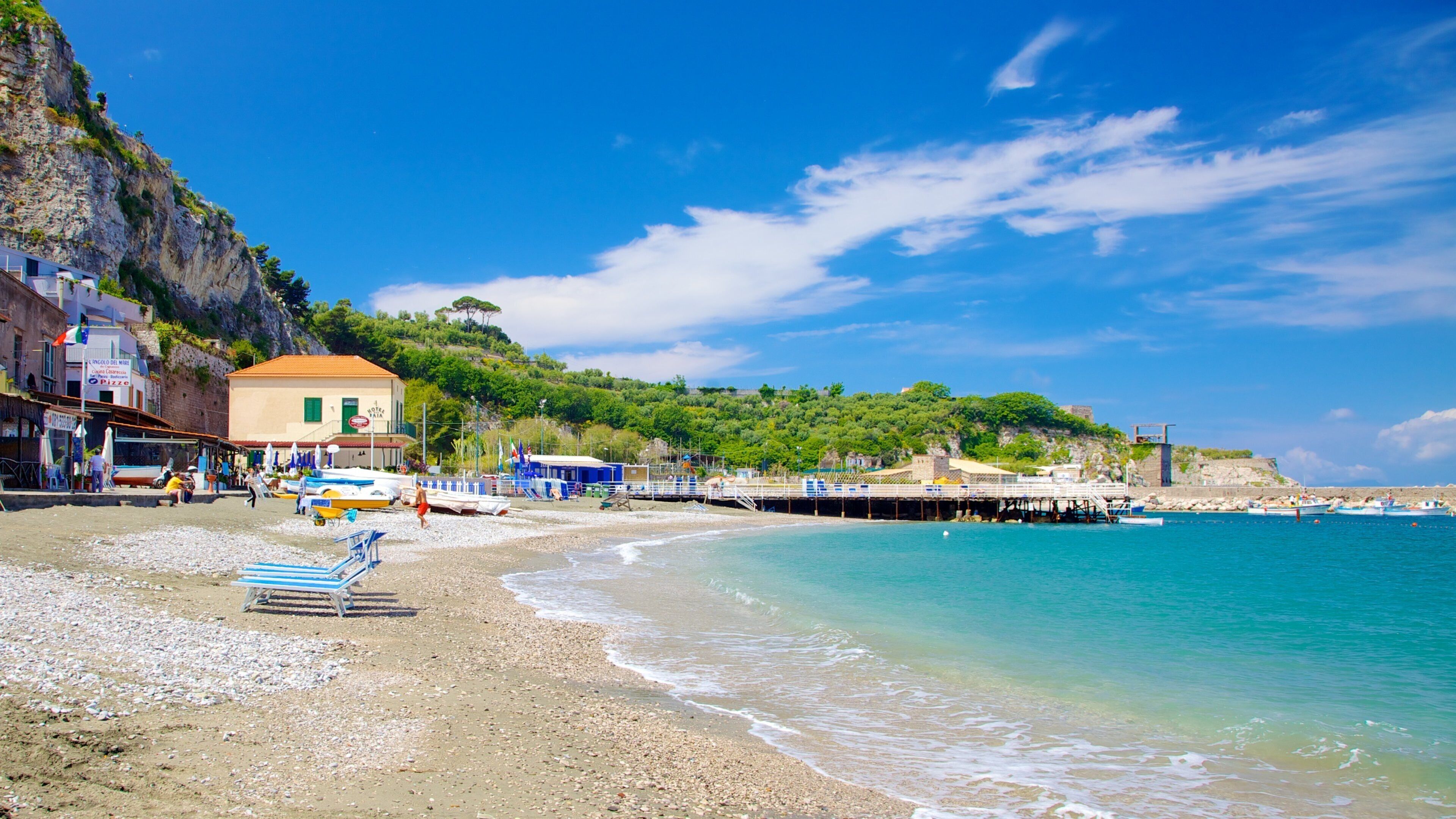 A beach with pebbles, a pier, boats, and buildings.