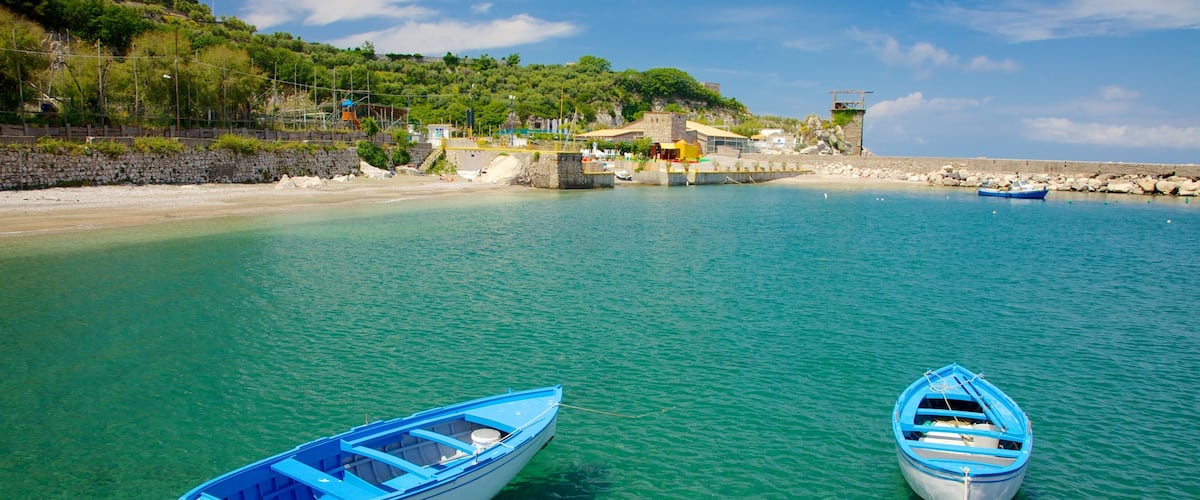 A coastal scene with two small boats tied to the shore, clear turquoise water, rocky beach, and a hill with buildings and trees in the background.