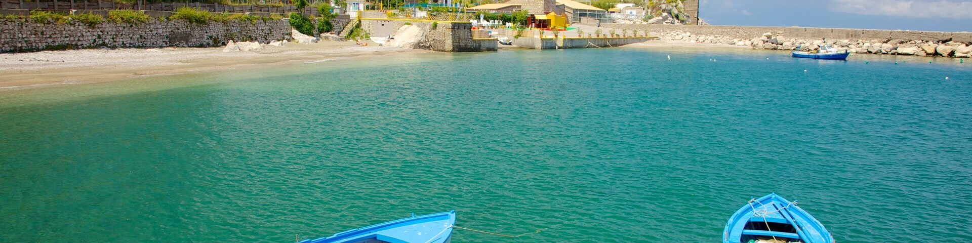 A coastal scene with two small boats tied to the shore, clear turquoise water, rocky beach, and a hill with buildings and trees in the background.