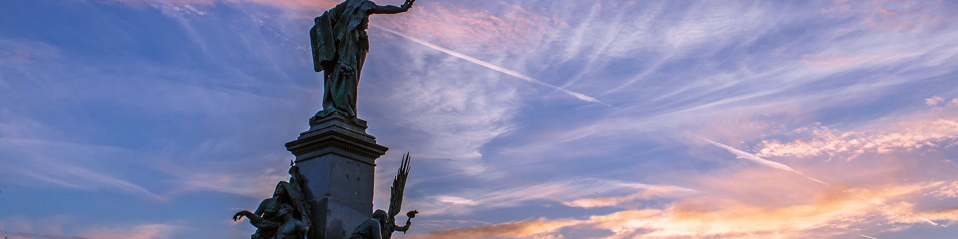 A statue of a figure with outstretched arms, a fountain, and an archway in a city square during sunset.