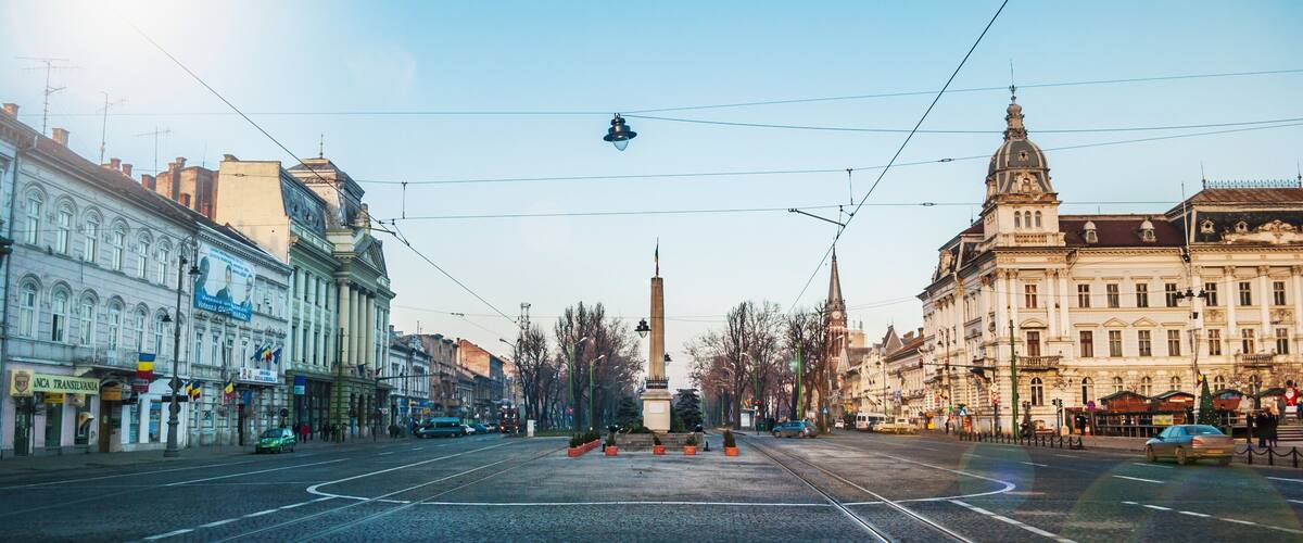 A city street with tram tracks, historic buildings, and a tall monument in the center.