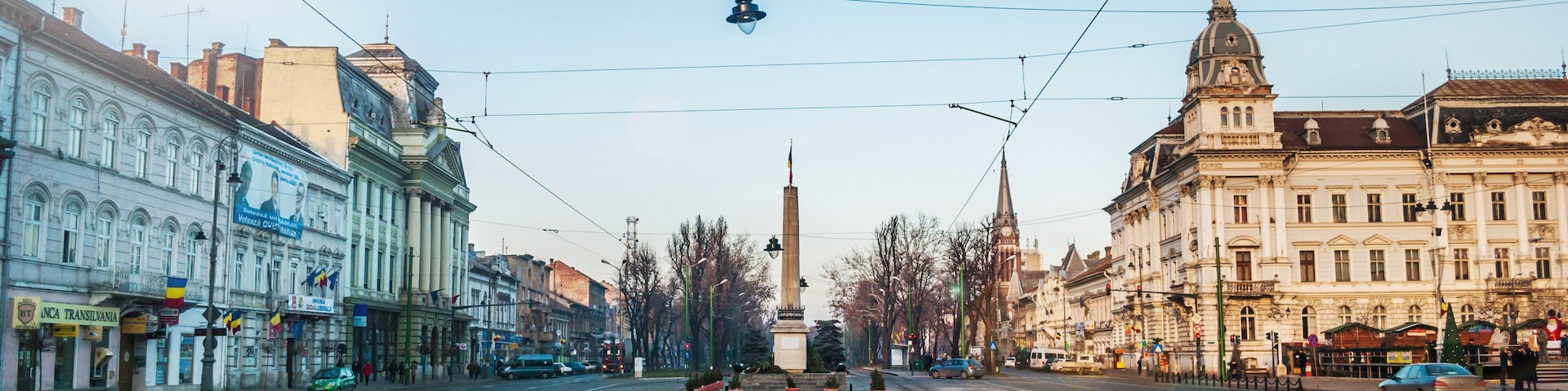A city street with tram tracks, historic buildings, and a tall monument in the center.