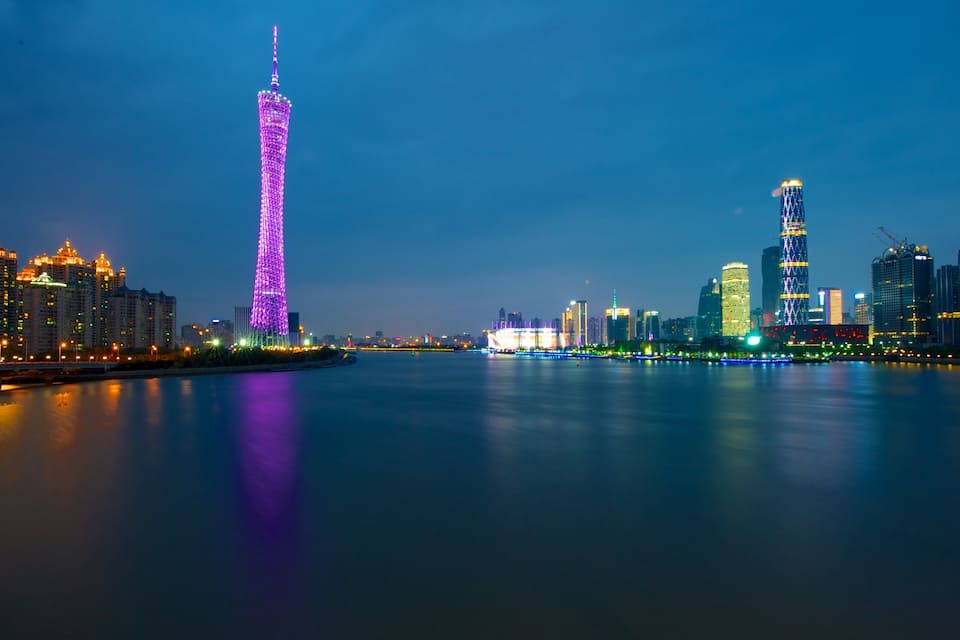 Horizonte urbano al anochecer con edificios iluminados y una torre destacada que se refleja en el agua.