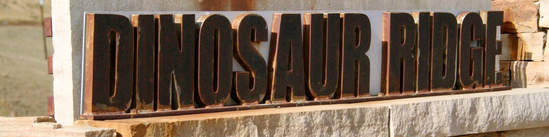 A sign reading "DINOSAUR RIDGE" mounted on a stone wall.