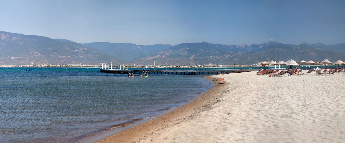 A beach with clear waters, a pier, and mountains in the background.
