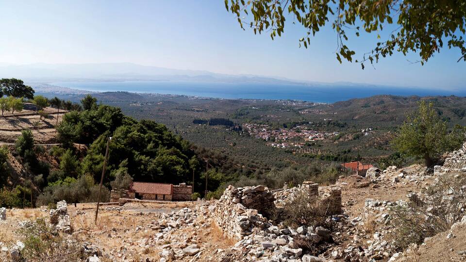 A coastal landscape with a stone house, ruins, and a view of the sea and distant mountains.