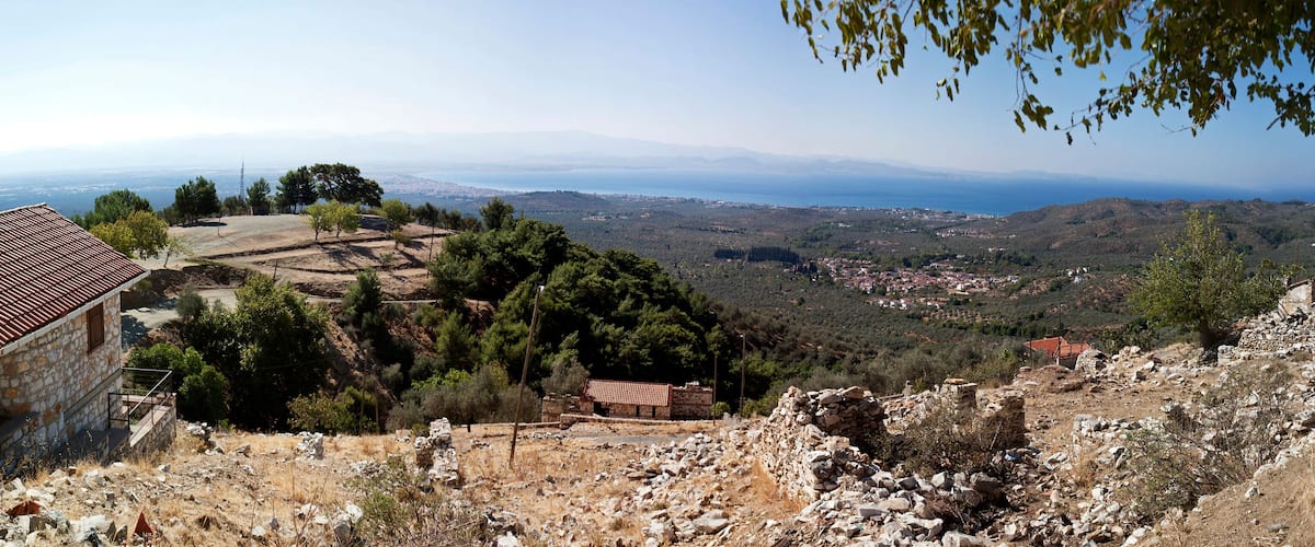 A coastal landscape with a stone house, ruins, and a view of the sea and distant mountains.