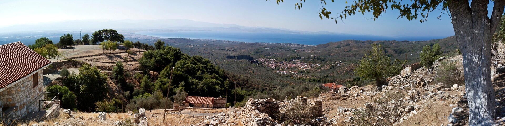 A coastal landscape with a stone house, ruins, and a view of the sea and distant mountains.