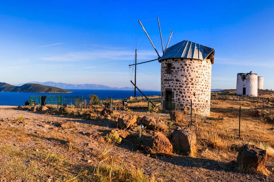 Un molí de vent en un turó, sota un cel blau clar i amb muntanyes al fons.