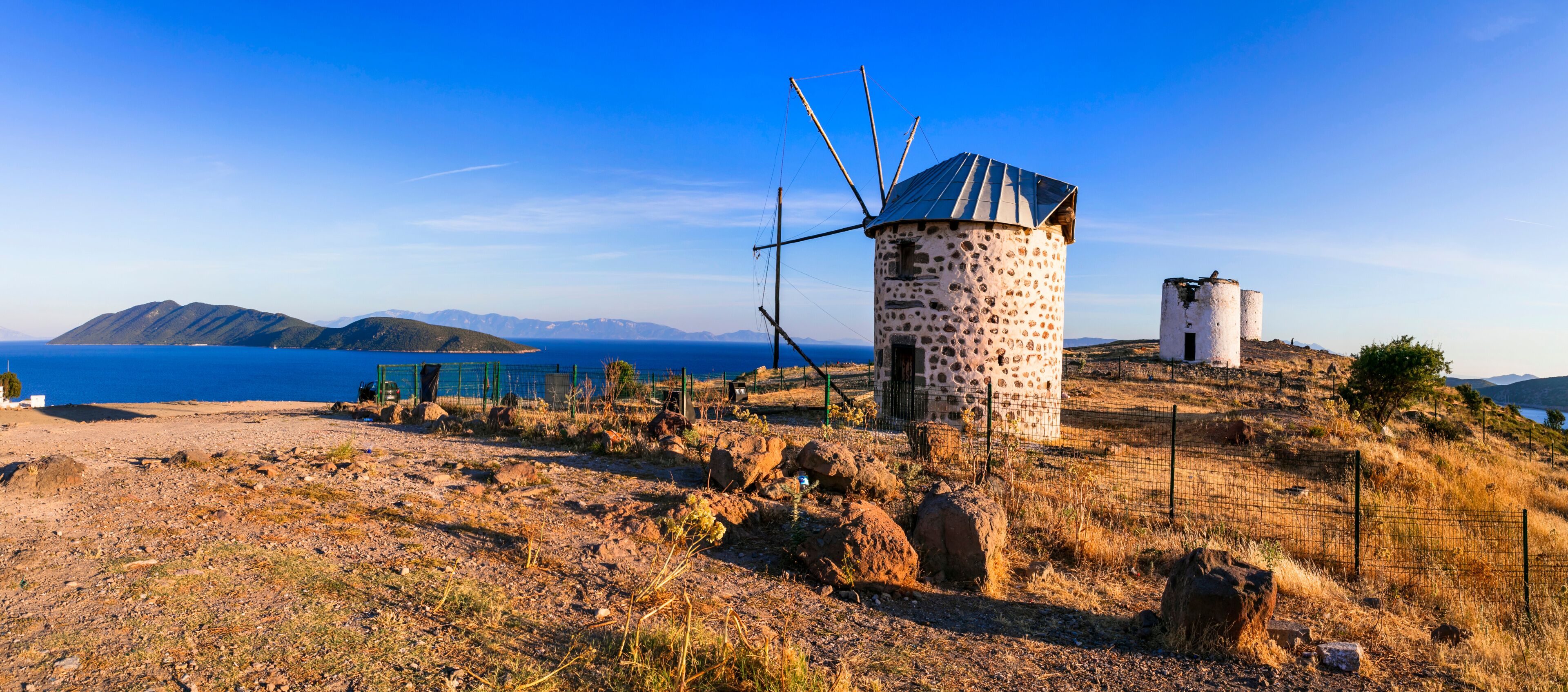 A windmill on a hillside with a clear blue sky and mountains in the background.