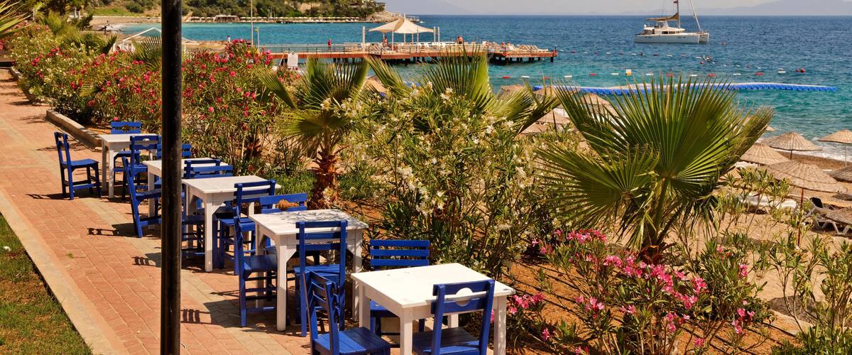 A coastal promenade with blue and white outdoor seating, a sailboat on the water, and a pier with umbrellas.