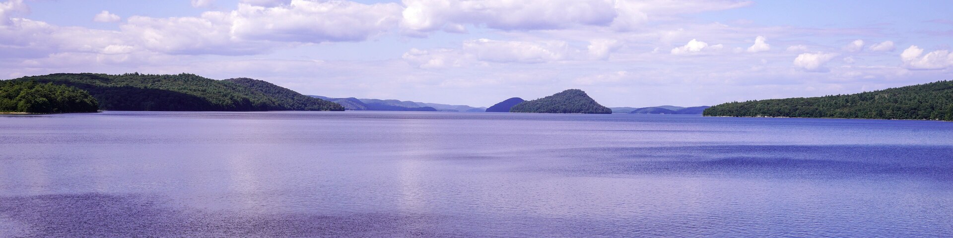 Un lago tranquilo con costas rocosas y colinas verdes distantes bajo un cielo parcialmente nublado.