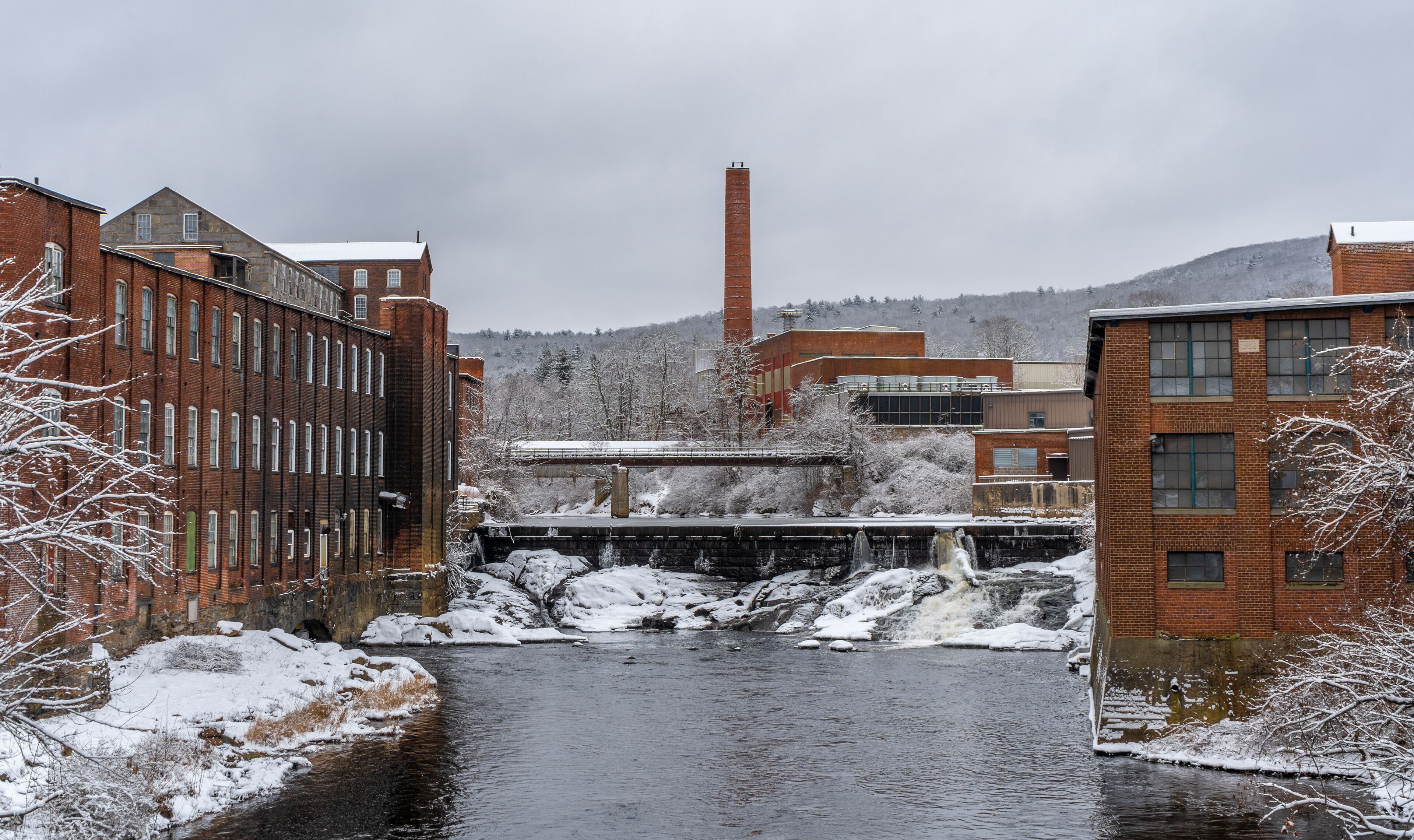 A snow-covered river flowing through a town with brick buildings and a tall chimney in the background.