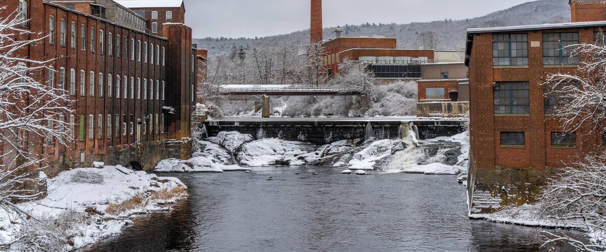A snow-covered river flowing through a town with brick buildings and a tall chimney in the background.