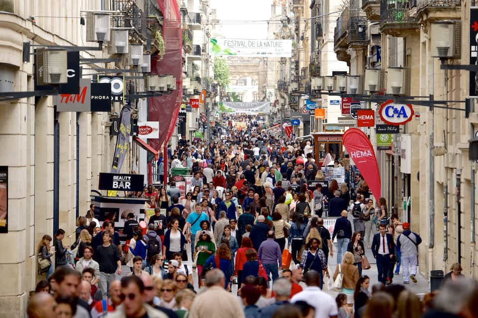 Une rue animée où l’on voit des gens se promener, un salon de beauté et divers panneaux, dont une affiche pour un festival.