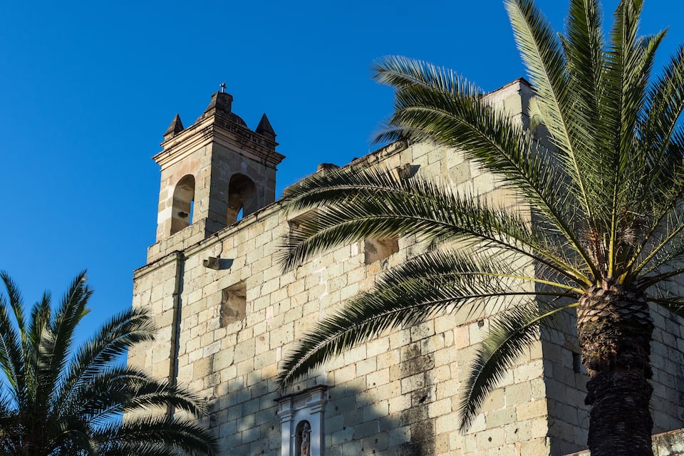 A historic stone building with a bell tower and palm trees.