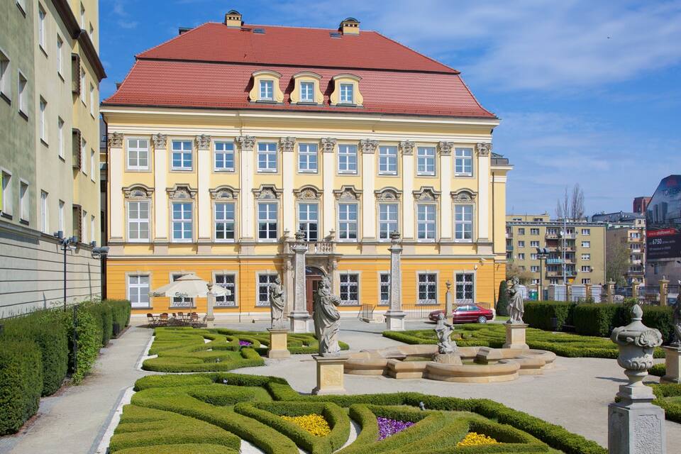 Un grand bâtiment jaune surmonté d’un toit rouge, entouré d’un jardin à la française agrémenté de statues et de haies taillées.