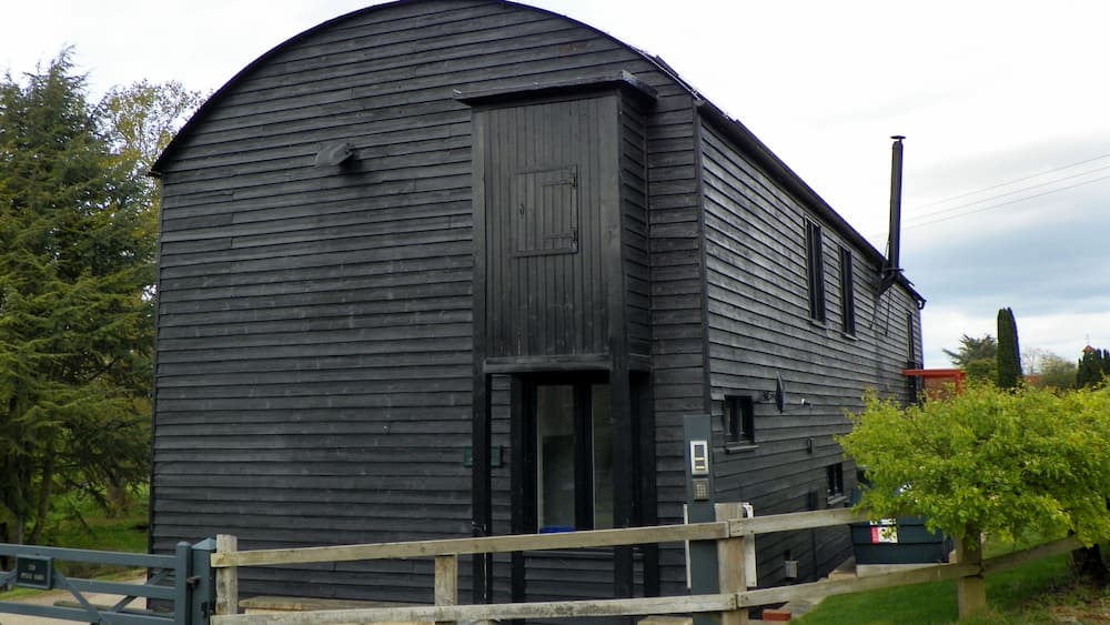 A large, dark wooden barn with a curved roof, surrounded by greenery and a wooden fence.