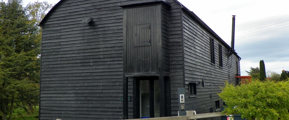 A large, dark wooden barn with a curved roof, surrounded by greenery and a wooden fence.