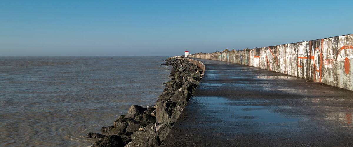 A coastal walkway with a rocky edge, a red and white lighthouse, and a concrete wall with graffiti.