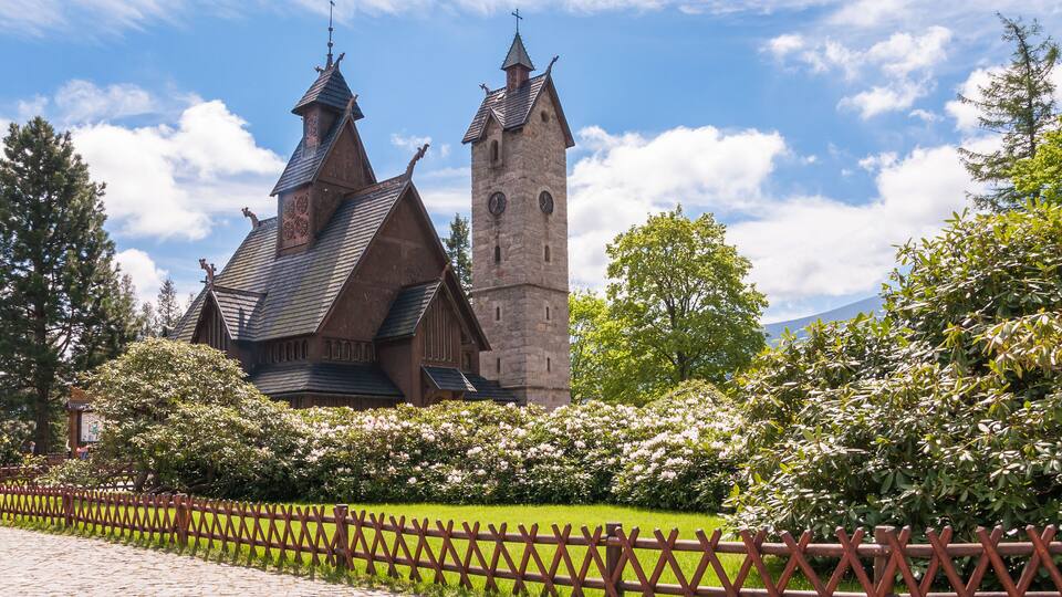 A historic building with a tall tower and steeply pitched roofs, surrounded by lush greenery and a wooden fence.